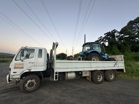 Stone & Rubble's truck transporting a blue tractor through a rural landscape with power lines.