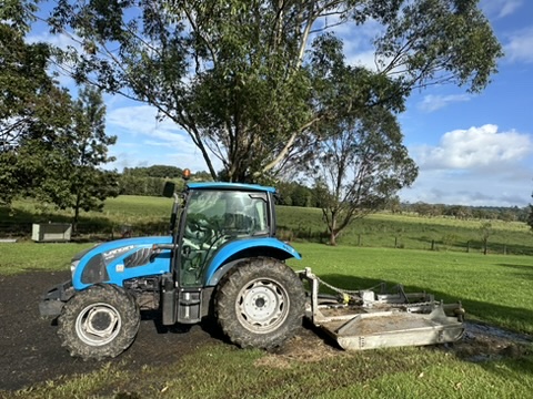 Blue tractor with cutting attachment in lush grass, surrounded by trees and rolling hills for Acreage Mowing Services.